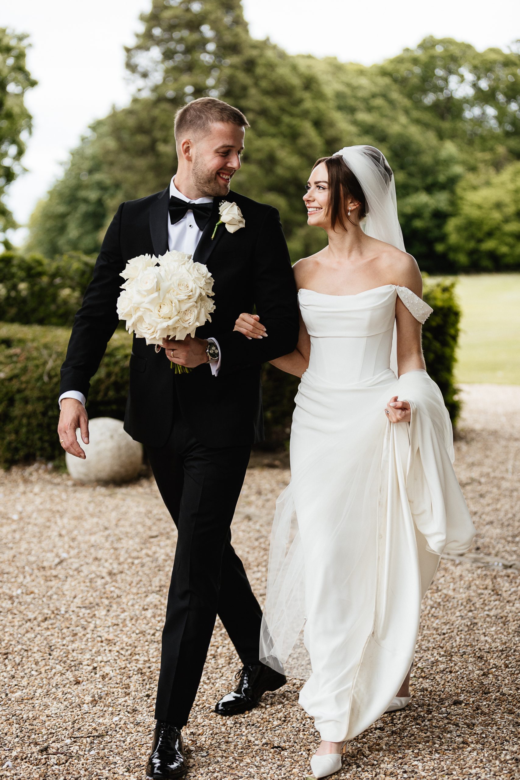Hertfordshire Wedding Photographer - Shrigley Photography & Films - SHRIGLEY Bride & Groom walking on a gravel path looking at each other and holding a bouquet of flowers - Editorial Wedding Photographer and Film In Hertfordshire | Shrigley Photography & Films