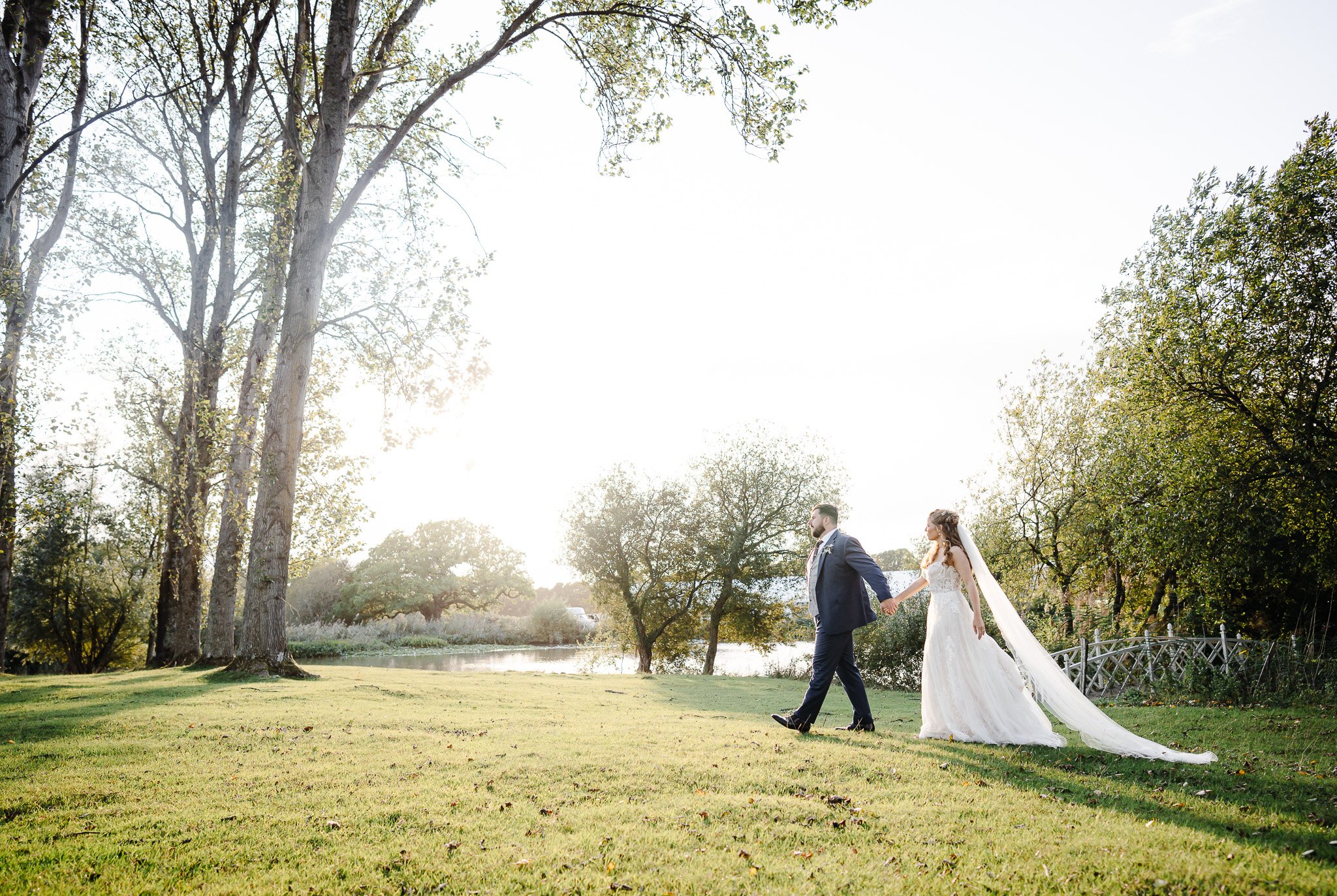 Hertfordshire Wedding Photographer - Shrigley Photography & Films - SHRIGLEY Bride & Groom walking holding hands by a lake on a green grass bank with lots of trees and evening sunshine - Wedding Photographer Hertfordshire -Shrigley Photo & Film