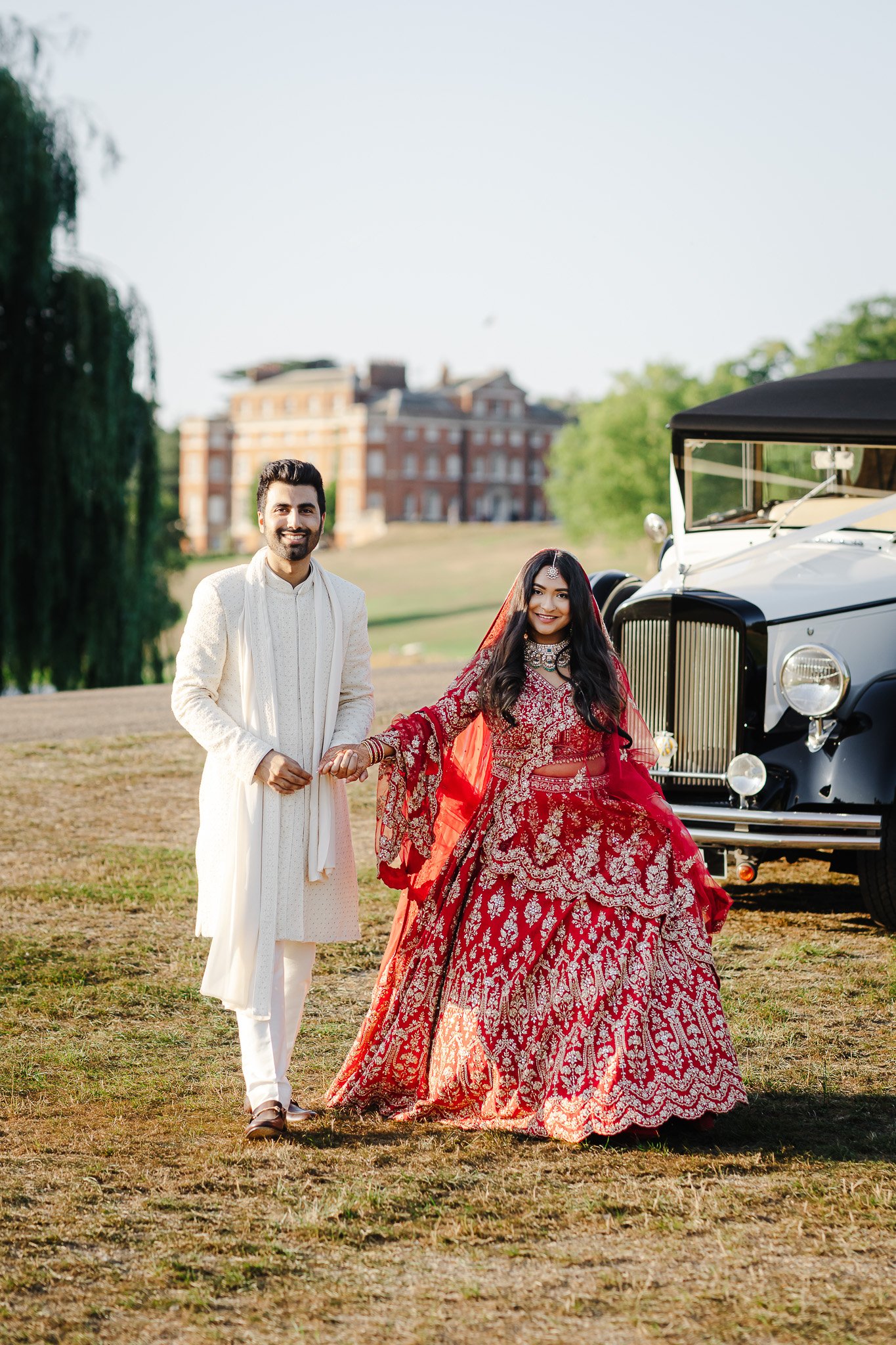 Hertfordshire Wedding Photographer & Videographer - Shrigley Photography - SHRIGLEY Man and woman walking towards the camera in indian wedding outfits, cream and red, with Brocket Hall in the background and classic car on the side- Hertfordshire Wedding Photographer & Videographer - Shrigley Photography