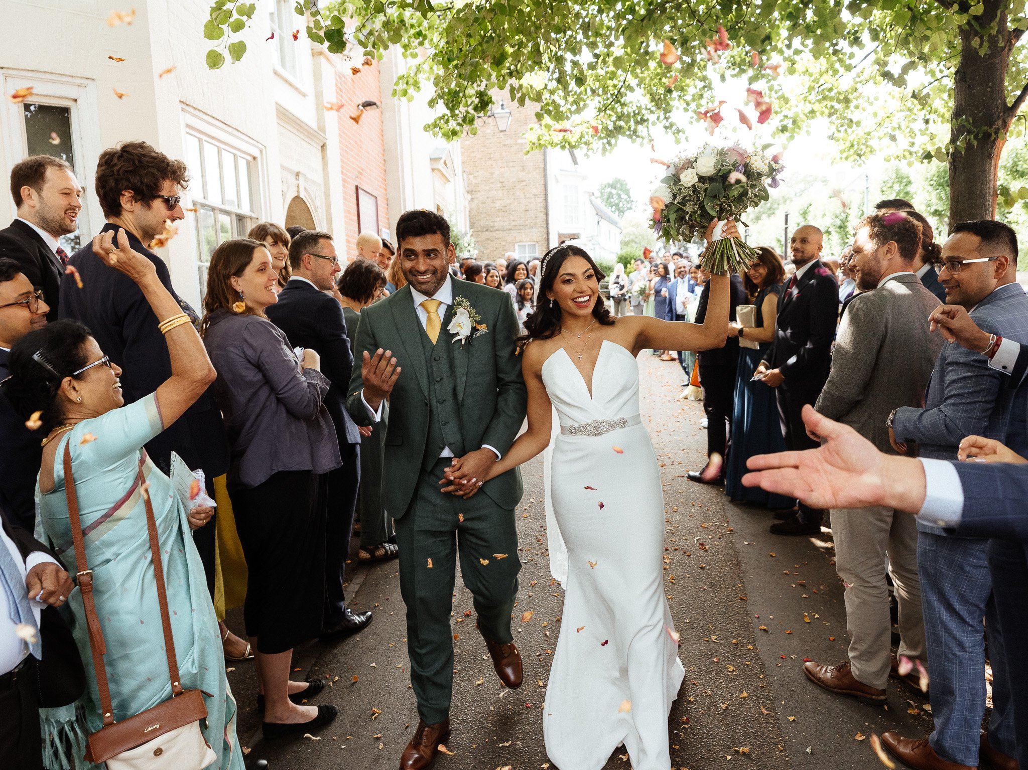 Hertfordshire Wedding Photographer - Shrigley Photography & Films - SHRIGLEY Bride & Groom walking in confetti tunnel with family waving and bride holding up her wedding bouquet - Quendon Hall Wedding | Shrigley Photography & Film